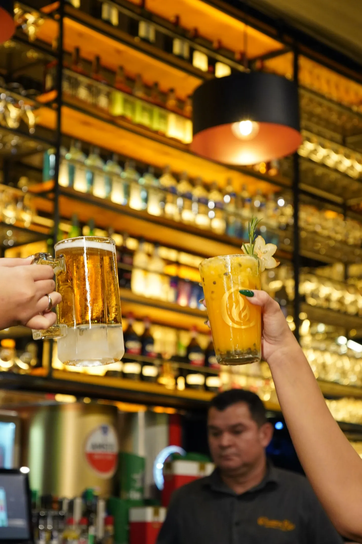 Brinde com chopp e drink de maracujá no bar do Chama Rio Choperia e Grill, em Copacabana, com estante de bebidas iluminada ao fundo